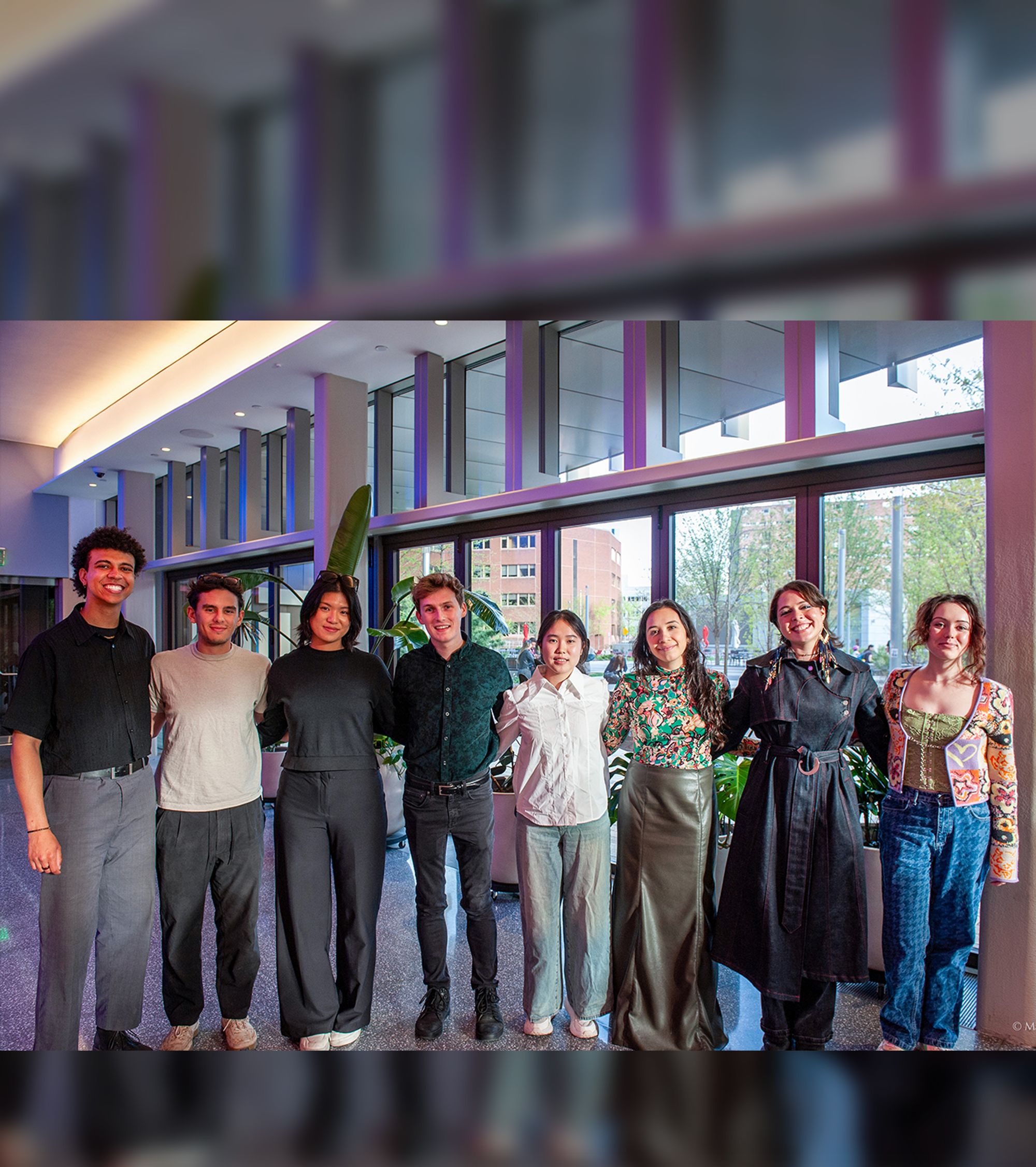 A group of eight young adults stands side by side in a bright, modern lobby with tall windows. They are smiling and facing the camera, dressed in a mix of casual and semi-formal outfits. Large plants and a view of trees and buildings are visible through the windows behind them.